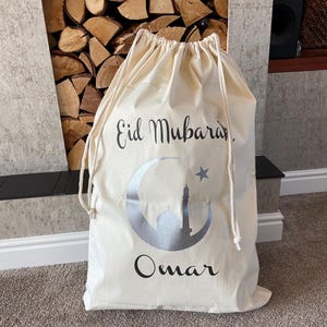May include: A white canvas drawstring bag with the text "Eid Mubarak" and a silver crescent moon with a star and a mosque inside. The name "Omar" is written below the moon.