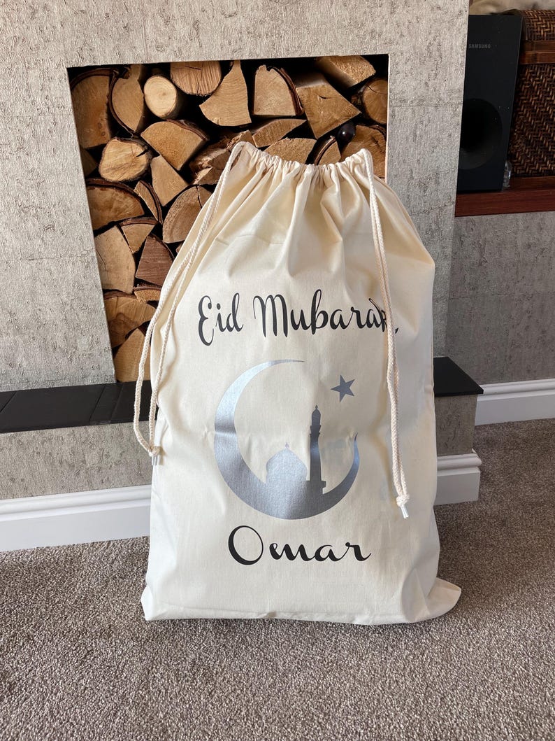 May include: A white canvas drawstring bag with the text "Eid Mubarak" and a silver crescent moon with a star and a mosque inside. The name "Omar" is written below the moon.