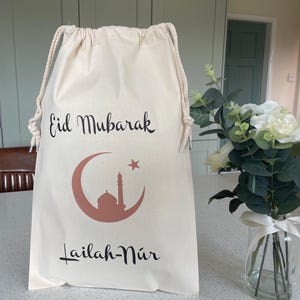 May include: A white drawstring bag with the text "Eid Mubarak" and a crescent moon with a star and a mosque inside. The text "Lailah-Nur" is below the moon.