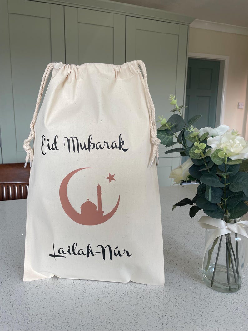 May include: A white drawstring bag with the text "Eid Mubarak" and a crescent moon with a star and a mosque inside. The text "Lailah-Nur" is below the moon.