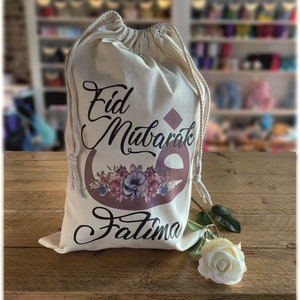 May include: A white drawstring bag with the text "Eid Mubarak Fatima" in black and brown lettering. The bag has a floral design with pink, blue, and white flowers. A white rose is lying on a wooden surface next to the bag.