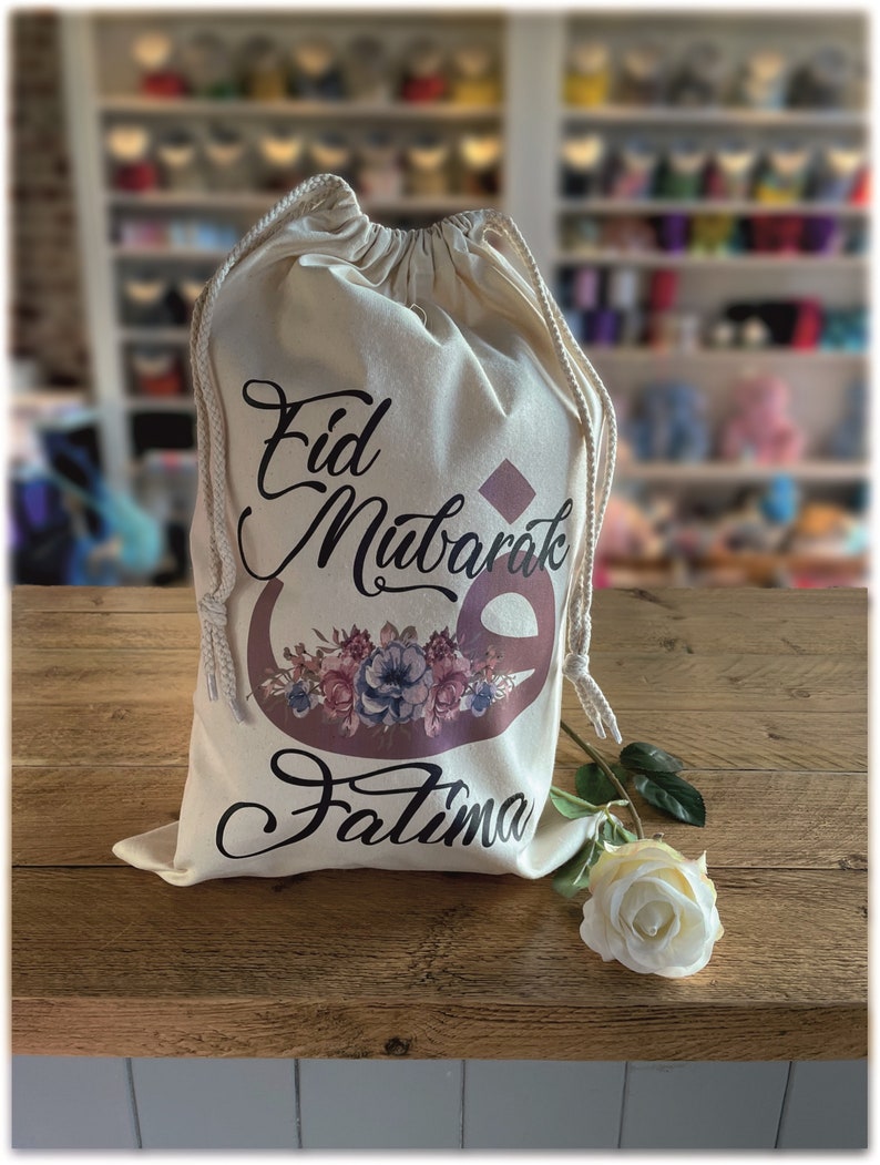 May include: A white drawstring bag with the text "Eid Mubarak Fatima" in black and brown lettering. The bag has a floral design with pink, blue, and white flowers. A white rose is lying on a wooden surface next to the bag.