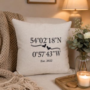 May include: A square, off-white linen-textured throw pillow featuring black printed text. The design displays the coordinates 54°02'18"N and 0°57'43"W separated by two small black hearts, with the text Est. 2022 printed underneath the coordinates.