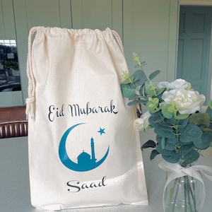 May include: A white cotton drawstring bag with the text "Eid Mubarak" and a blue crescent moon and mosque design. The bag also has the name "Saaad" printed on it.