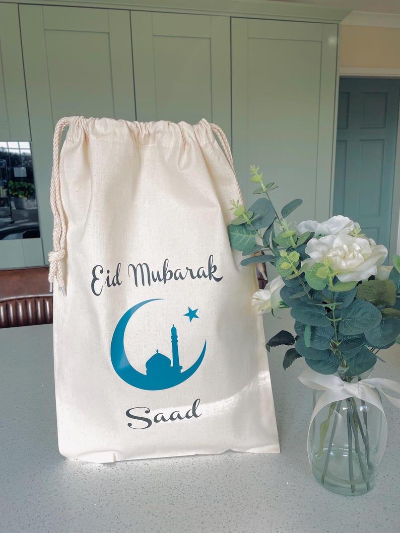 May include: A white cotton drawstring bag with the text "Eid Mubarak" and a blue crescent moon and mosque design. The bag also has the name "Saaad" printed on it.