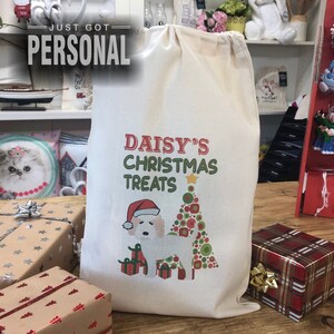 Könnte beinhalten: Eine weiße Stofftasche mit einem roten und grünen Weihnachtsmotiv. Die Tasche zeigt einen weißen Hund mit einem Weihnachtsmann-Hut, einen Weihnachtsbaum und den Text "Daisy's Christmas Treats".