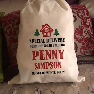 May include: A white canvas Santa sack with red text that reads "SPECIAL DELIVERY FROM THE NORTH POLE FOR PENNY SIMPSON DO NOT OPEN UNTIL DEC 25". The sack has a green and red illustration of a house with a snowflake and two Christmas trees.