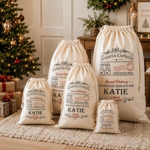 May include: A set of five off-white drawstring canvas bags in varying sizes, each printed with black and red text reading Reindeer Express, Special Delivery, Checked Once Twice, North Pole Mail, and the name Katie in large letters.
