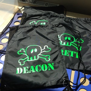 Könnte beinhalten: Schwarze Kordelzugbeutel mit einem neongrünen Totenkopf- und gekreuzten Knochen-Design. Das Wort "DEACON" ist unter dem Totenkopf auf einer Tasche aufgedruckt, und "BRETT" auf einer anderen. Die Taschen haben schwarze Kordelzüge.