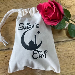 May include: A white drawstring bag with a black crescent moon, star, and mosque design. The bag is decorated with the text "Saira's Eid" in black.