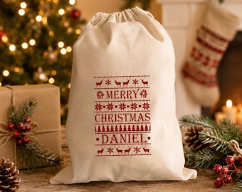 May include: A cream-colored drawstring gift bag featuring a red printed design with rows of reindeer, snowflakes, and pine trees. The text "MERRY CHRISTMAS" and the name "DANIEL" are printed in red in the center of the bag.