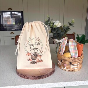May include: A beige drawstring bag with a woodland animal design and the text "Happy Easter Sofia". Next to it is a woven basket shaped like a bunny, filled with Easter eggs and carrots.