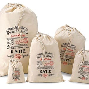 May include: A set of five off-white canvas drawstring bags in varying sizes, each printed with black and red text reading Reindeer Express, Special Delivery, North Pole Mail, and the name Katie, featuring check boxes and decorative flourishes.