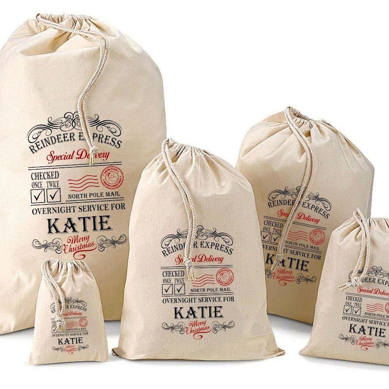 May include: A set of five off-white canvas drawstring bags in varying sizes, each printed with black and red text reading Reindeer Express, Special Delivery, North Pole Mail, and the name Katie, featuring check boxes and decorative flourishes.