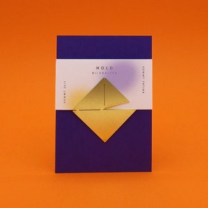 May include: A gold-colored metal picture holder with the text "HOLD BILDHALTER" and "KOMMT PATINA KOMMT ZEIT" on a white background. The holder is on a purple background.