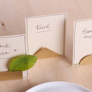 May include: Three gold metal place card holders with a triangular and semi-circular design. The place cards are white with black ink and say "Kurt", "Hans", and "Tante Lisa".