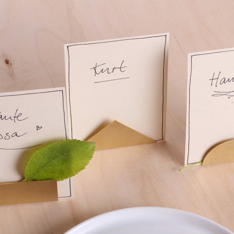 May include: Three gold metal place card holders with a triangular and semi-circular design. The place cards are white with black ink and say "Kurt", "Hans", and "Tante Lisa".
