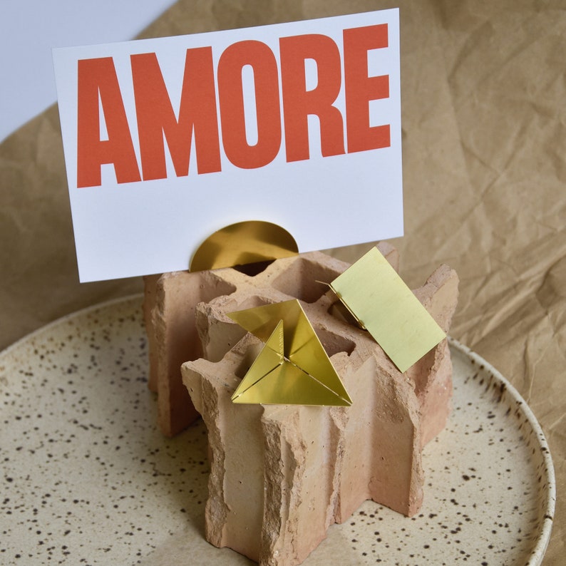 May include: A terracotta brick-shaped card holder with a gold metal top. The card holder is holding a white card with the word "AMORE" printed in red letters. There are two gold metal origami shapes on the holder.