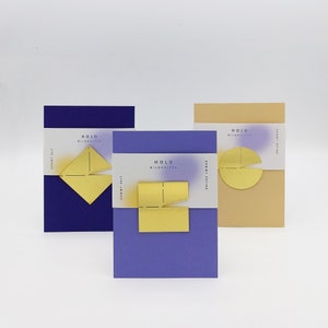 May include: Three different colored cards with the text "HOLD BILDHALTER" and a geometric shape in gold. The cards are purple, blue, and beige.
