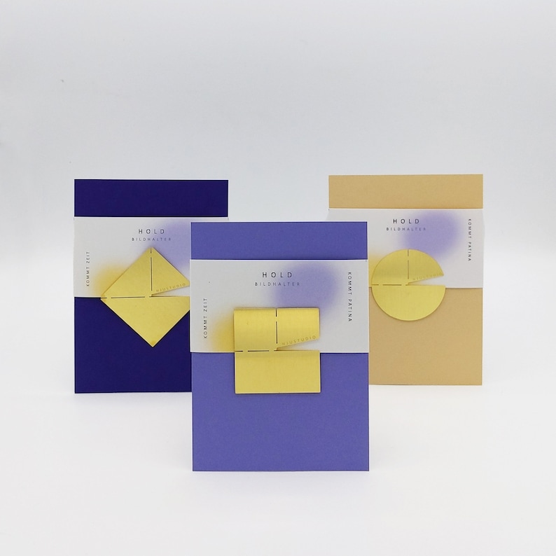 May include: Three different colored cards with the text "HOLD BILDHALTER" and a geometric shape in gold. The cards are purple, blue, and beige.