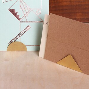 May include: Two brass paperweights, one shaped like a half-circle and the other like a triangle, are used to hold down a brown envelope and a light blue card with a geometric design.
