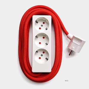 Puede incluir: Una regleta blanca de tres enchufes con un cable cubierto de tela roja. El cable está enrollado alrededor de la regleta. El texto "RED" es visible debajo de la regleta.
