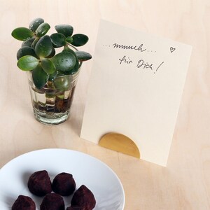 May include: A small plant in a glass container sits on a wooden surface next to a white card with the handwritten message "... mmh... f&uuml;r Dich!" and a small heart. A gold metal stand holds the card upright.