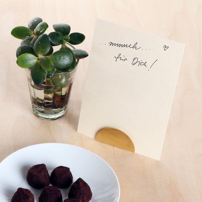 May include: A small plant in a glass container sits on a wooden surface next to a white card with the handwritten message "... mmh... f&uuml;r Dich!" and a small heart. A gold metal stand holds the card upright.