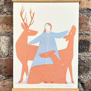 Könnte beinhalten: Gerahmter Kunstdruck mit einer Frau in einem blauen Kleid mit drei orangefarbenen Hirschen. Das Kunstwerk hat einen minimalistischen Stil mit hellem Hintergrund und wird mit einem Holzrahmen präsentiert. Der Druck hängt an einer Backsteinmauer.