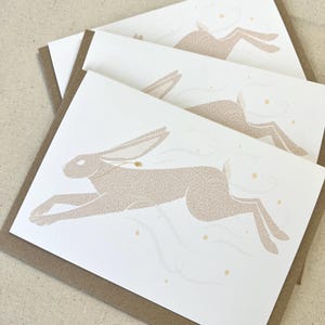 May include: Three greeting cards featuring a running hare illustration in beige on white cardstock. The cards have a brown border and are stacked. The hare is depicted with flowing lines and small gold stars.