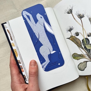 May include: A blue bookmark featuring a white rabbit illustration with a starry night sky design. The bookmark is inserted into a book with botanical illustrations. The website address "www.abbielub.com" is printed on the bookmark.