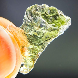 Può includere: Una tectite moldavite verde oliva traslucida con una superficie irregolare e altamente strutturata caratterizzata da piccole fosse, creste e una distinta rientranza circolare. Il minerale mostra varie sfumature di verde e marrone con una lucentezza vitrea.