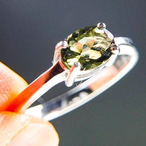 May include: A silver ring with an oval, olive-green gemstone. The ring has a thin, polished band and four prongs securing the gemstone. The ring is held by a finger against a dark background. The gemstone is a light green colour.