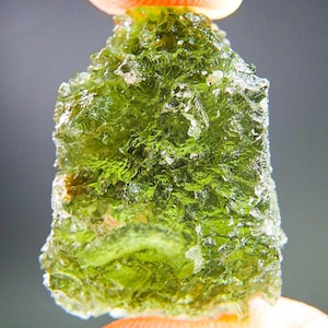 Peut inclure: Gros plan sur une pierre verte translucide, rugueuse et de forme irrégulière. La pierre a une surface texturée avec un aspect mousseux et est tenue entre deux doigts. La couleur est un vert naturel vibrant avec des zones plus claires, presque blanches.