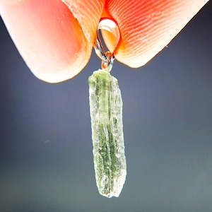 Puede incluir: Un colgante de cristal bruto de color verde claro translúcido con una superficie rugosa y texturizada. La piedra alargada e irregular está sujeta a una anilla de metal plateado pulido en la parte superior para usarla como dije.