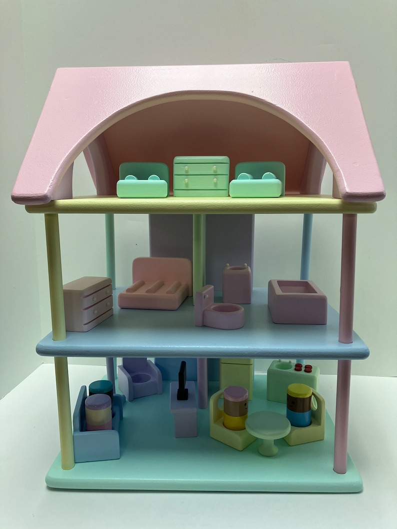 May include: A pastel-colored wooden dollhouse with two stories and a pink roof. The dollhouse is furnished with miniature furniture, including sofas, a bed, a dresser, a toilet, a bathtub, and a table. The color scheme includes pink, blue, green, and yellow.