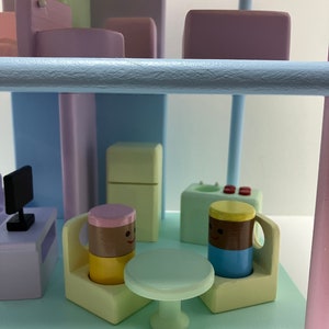 May include: A whimsical wooden miniature scene with pastel colors. Features a small table, chairs, and smiling mug-shaped figures. Includes a miniature television and other building blocks, creating a playful, imaginative setting.