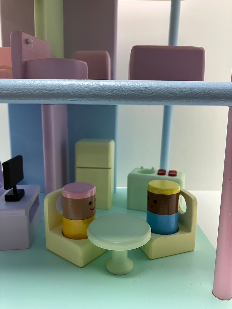 May include: A whimsical wooden miniature scene with pastel colors. Features a small table, chairs, and smiling mug-shaped figures. Includes a miniature television and other building blocks, creating a playful, imaginative setting.