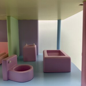 May include: A pastel-colored wooden dollhouse bathroom set. The set includes a pink toilet, a pink bathtub, and a pink sink. The walls are painted in pastel green and purple. The floor is light blue.