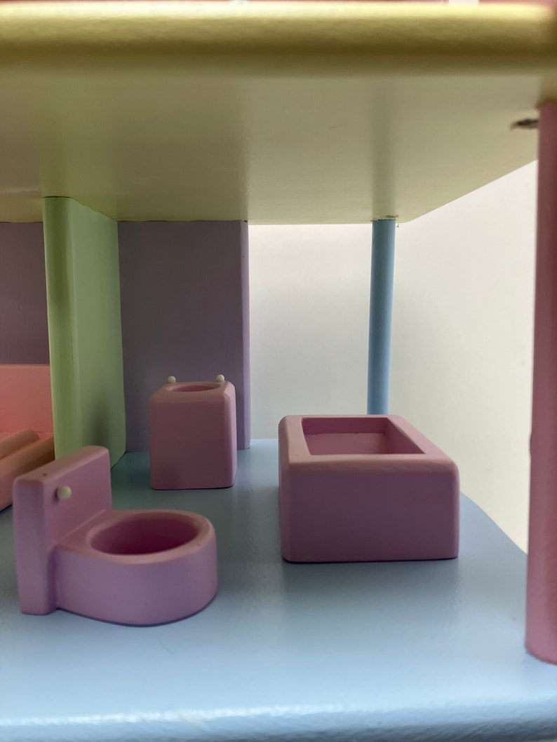 May include: A pastel-colored wooden dollhouse bathroom set. The set includes a pink toilet, a pink bathtub, and a pink sink. The walls are painted in pastel green and purple. The floor is light blue.