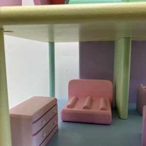 May include: A pastel-colored dollhouse interior with a light blue floor and walls in shades of pink, purple, and green. The miniature furniture includes a pink dresser, a pink sofa, and other small pieces. The dollhouse has a whimsical, playful design.