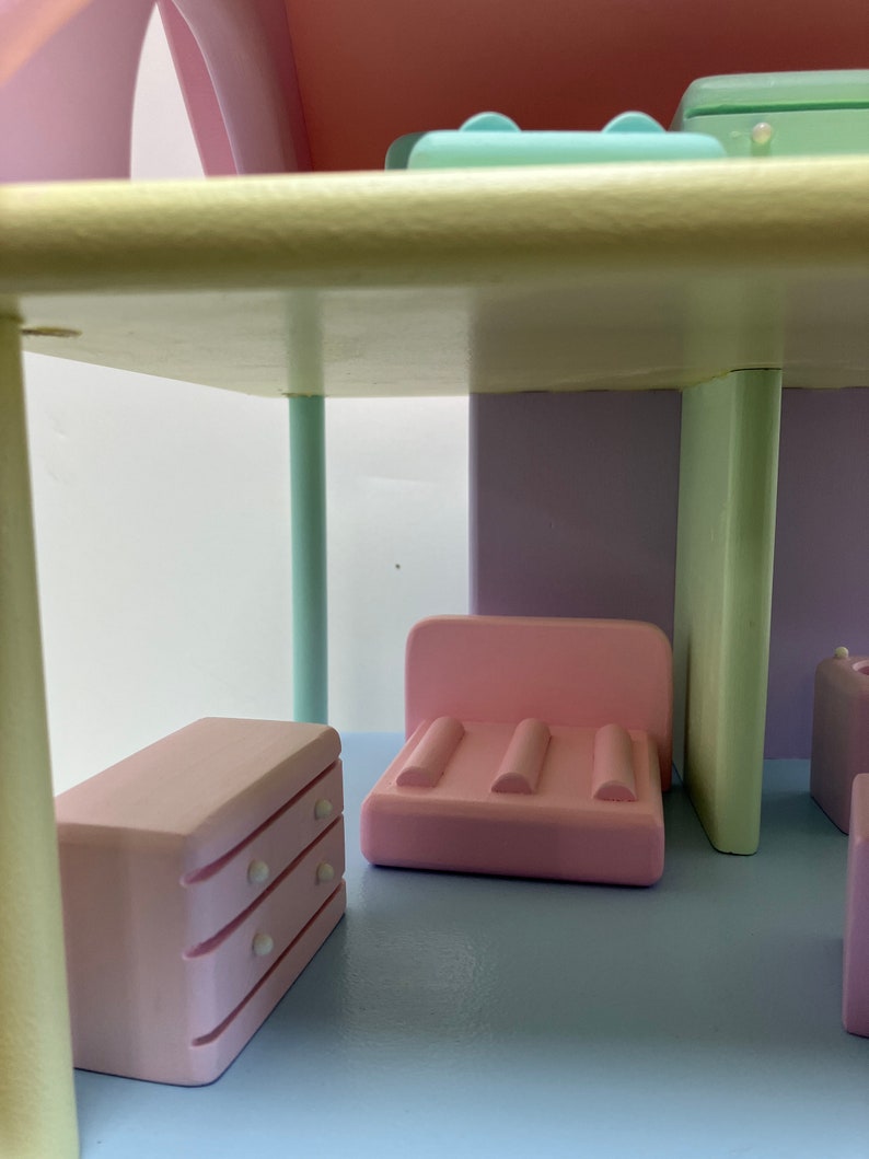 May include: A pastel-colored dollhouse interior with a light blue floor and walls in shades of pink, purple, and green. The miniature furniture includes a pink dresser, a pink sofa, and other small pieces. The dollhouse has a whimsical, playful design.