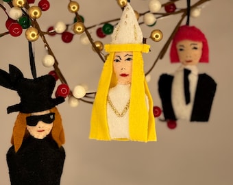 Puede incluir: Tres adornos de fieltro colgados de una rama. El adorno de la izquierda es una figura con sombrero negro y gafas de sol. El central es una figura con un sombrero blanco alto y tocado amarillo con una cadena dorada. El de la derecha es una figura con pelo rosa.