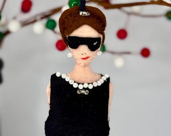 Puede incluir: Un adorno de fieltro que representa una figura con cabello castaño, que lleva gafas de sol negras grandes, una blusa negra sin mangas y un collar de cuentas blancas tipo perla. La figura tiene pequeños pendientes blancos y labios rojos, suspendida por una cinta negra.