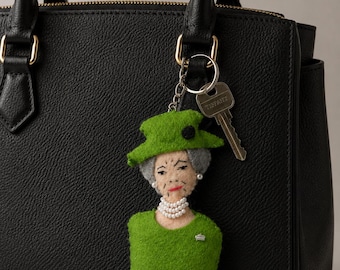 Puede incluir: Llavero de fieltro verde con la forma de una mujer con sombrero y vestido verdes, con collar de perlas y un broche plateado. Colgando de un bolso de mano de cuero negro, con una llave.