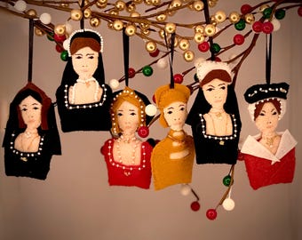 Puede incluir: Seis adornos de fieltro que representan figuras históricas, cada una con un tocado y atuendo únicos. Las figuras están hechas de tela con rasgos faciales bordados y detalles de cuentas, suspendidas por cintas negras de una rama con bayas doradas y rojas.