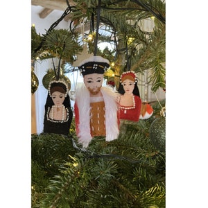 Puede incluir: Tres adornos de fieltro colgados de un &aacute;rbol. El adorno central es una figura con gorro negro, t&uacute;nica roja y ribete de piel sint&eacute;tica blanca. El adorno izquierdo es una figura con capucha negra y perlas blancas. El derecho es una figura con vestido rojo.