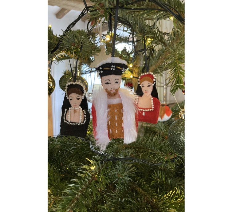 Puede incluir: Tres adornos de fieltro colgados de un &aacute;rbol. El adorno central es una figura con gorro negro, t&uacute;nica roja y ribete de piel sint&eacute;tica blanca. El adorno izquierdo es una figura con capucha negra y perlas blancas. El derecho es una figura con vestido rojo.