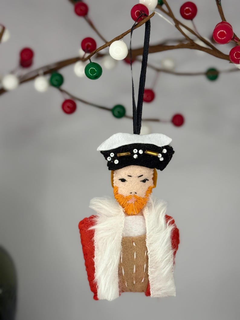 Puede incluir: Un adorno de fieltro que representa una figura con barba naranja, con un sombrero negro y blanco con detalles dorados, un abrigo rojo con ribete de piel sint&eacute;tica blanca y un chaleco color canela con costuras blancas.