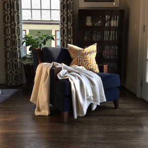 May include: A navy blue armchair with a white throw blanket and a yellow and brown geometric patterned pillow. The chair is in a living room with a wooden floor and a bookcase in the background.